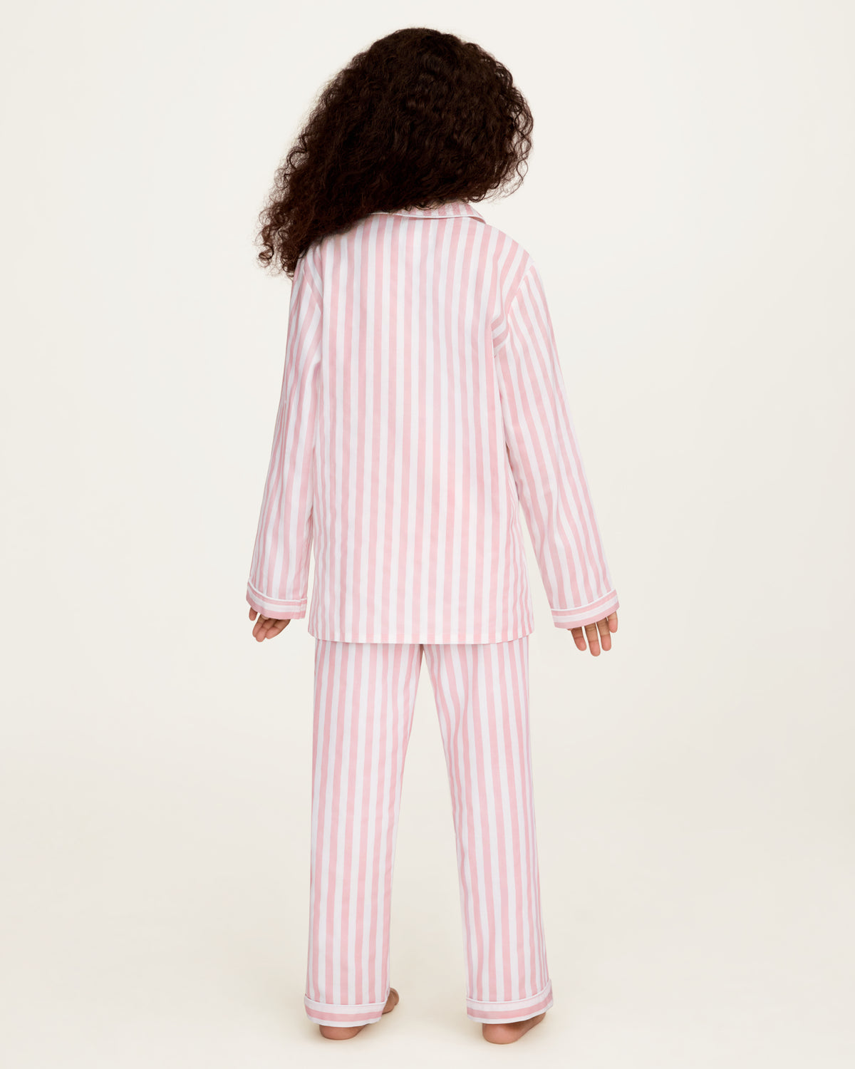 A child shown from the back wearing a two-piece pajama set with vertical pink and white stripes, standing against a plain off-white studio background.