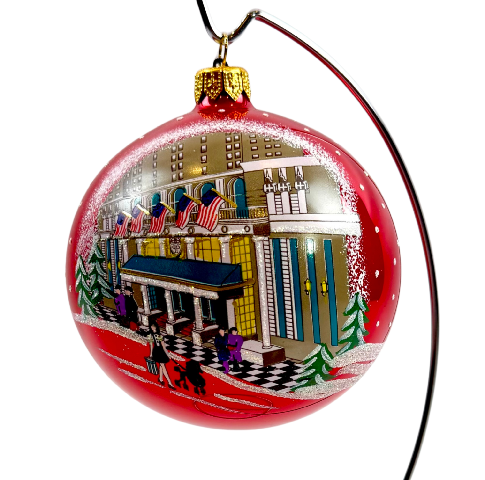 A red glass Christmas ornament featuring a hand-painted winter scene of a grand hotel facade with snow, trees, and pedestrians, suspended by a black string with a gold cap.