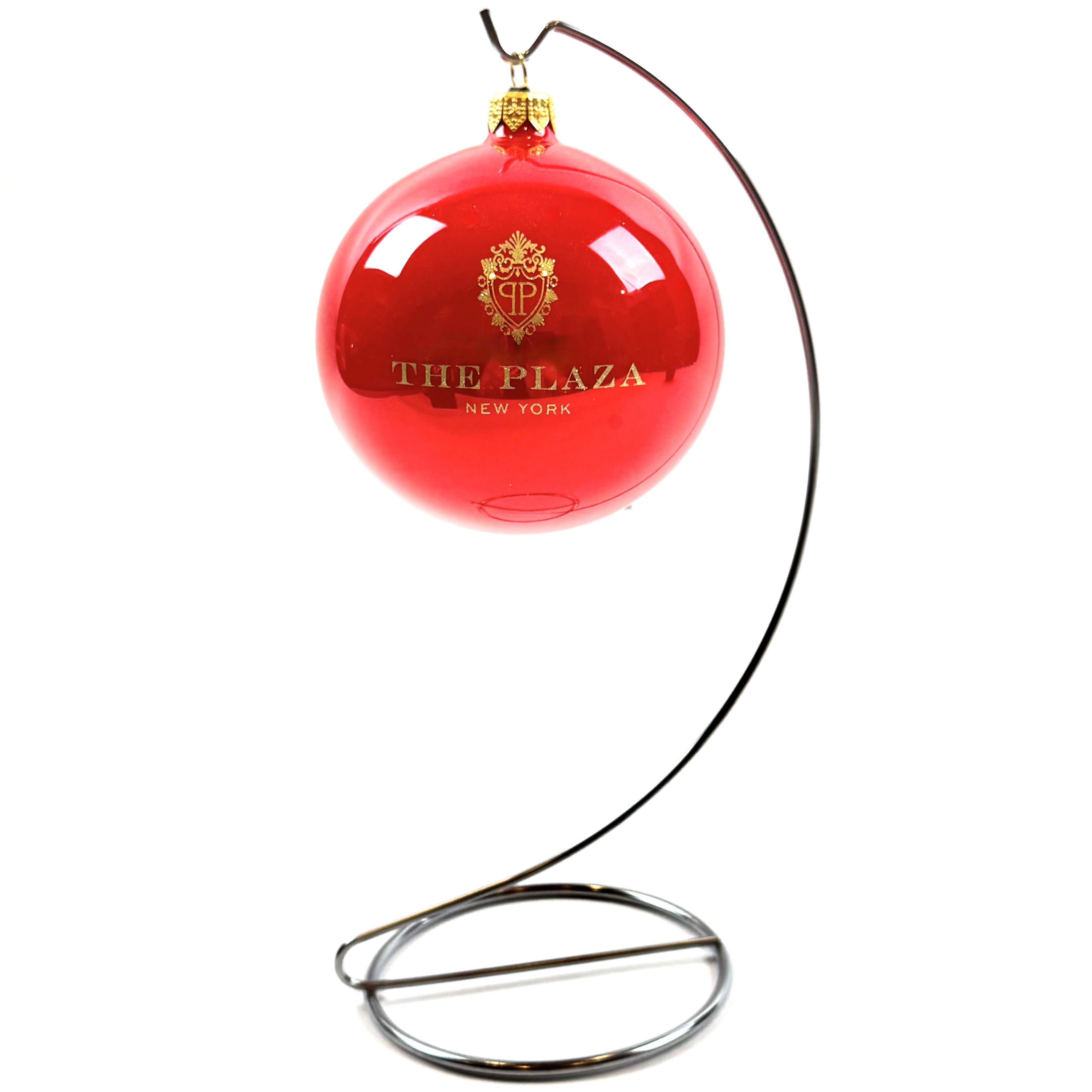 A glossy red Christmas ornament featuring the gold 'THE PLAZA NEW YORK' logo, suspended from a curved black metal stand against a white background.