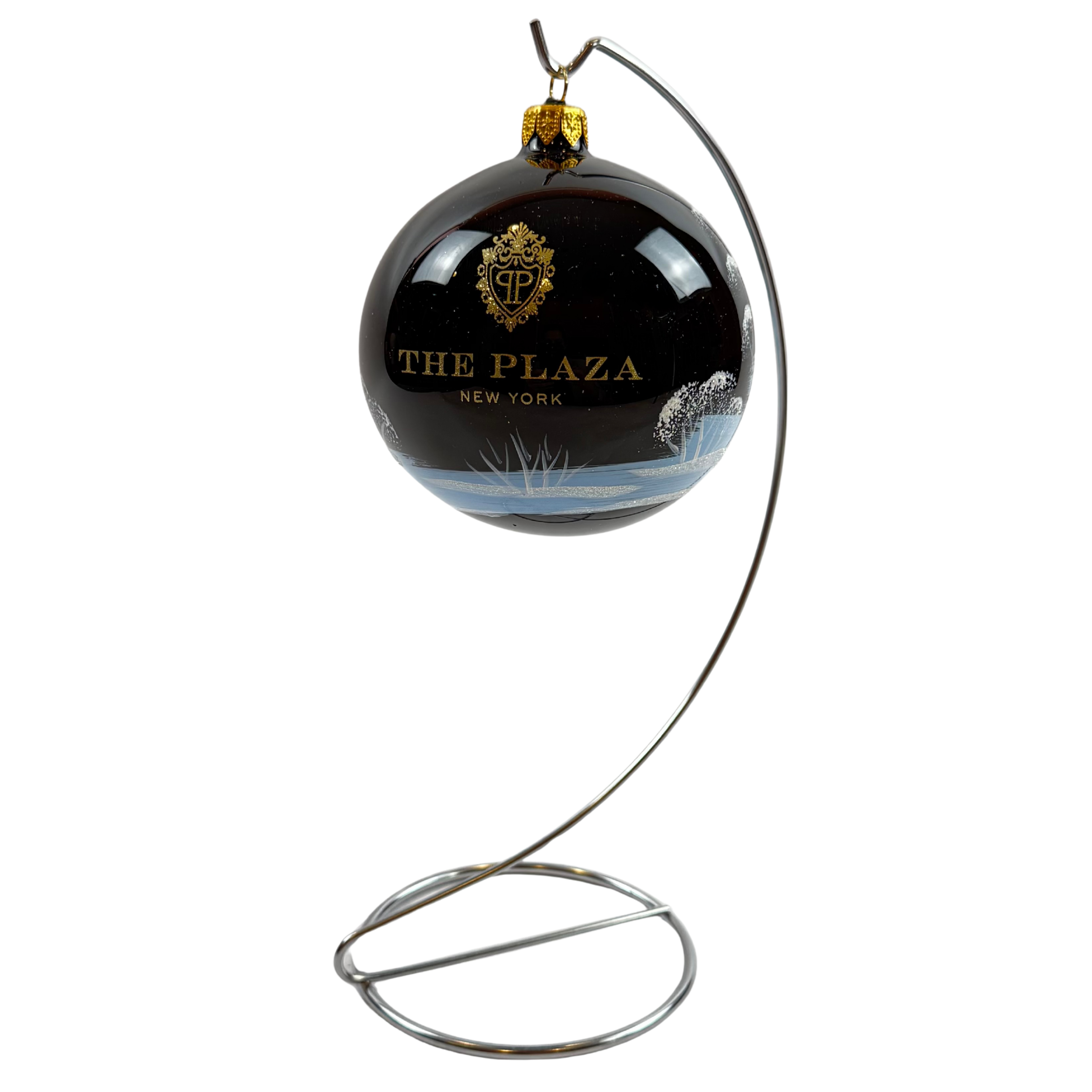 A spherical black Christmas ornament featuring a gold crest and the text 'THE PLAZA NEW YORK', suspended from a curved silver metal stand against a plain white background.