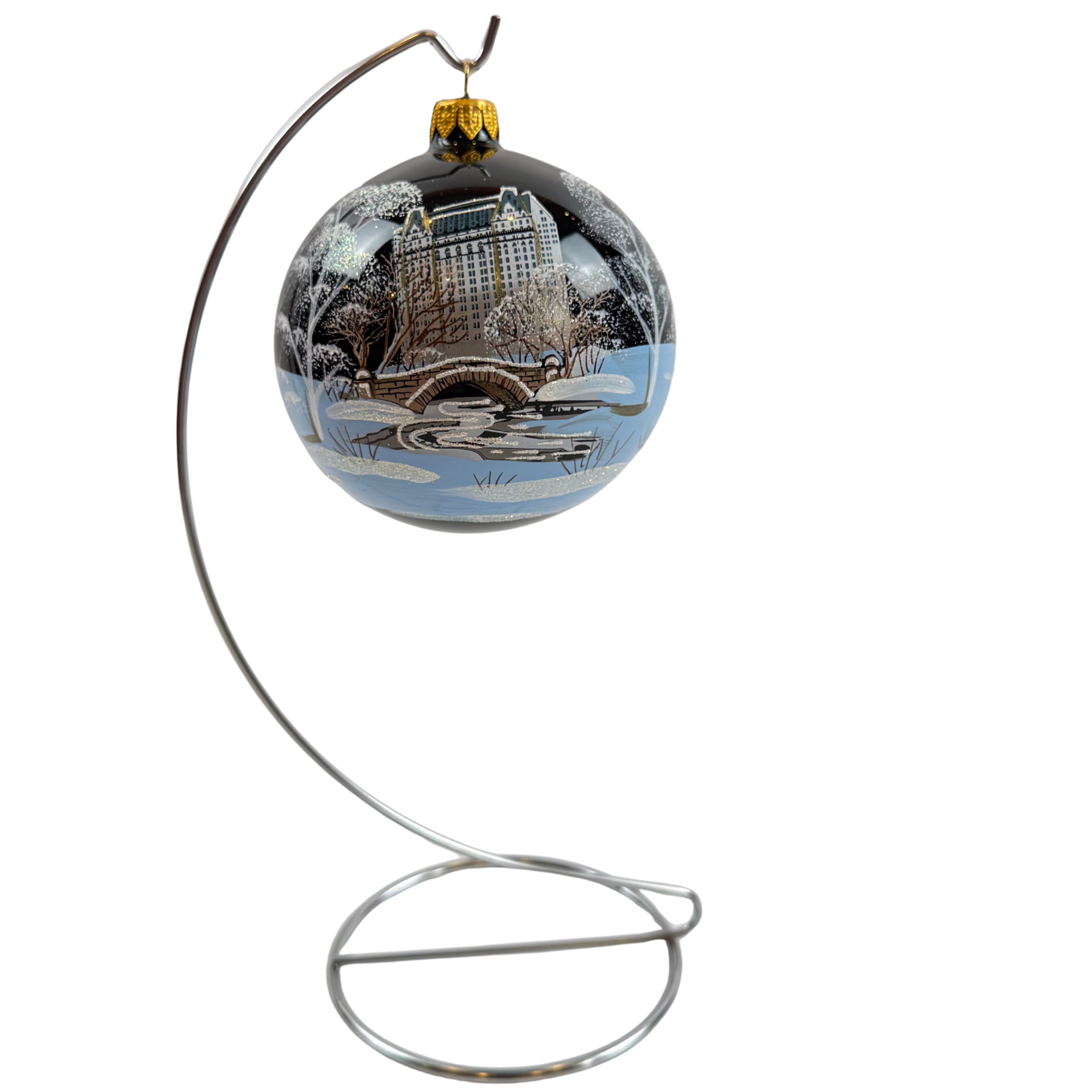 A decorative glass Christmas ornament with a winter cityscape print, hanging from a curved silver metal stand against a plain white background.