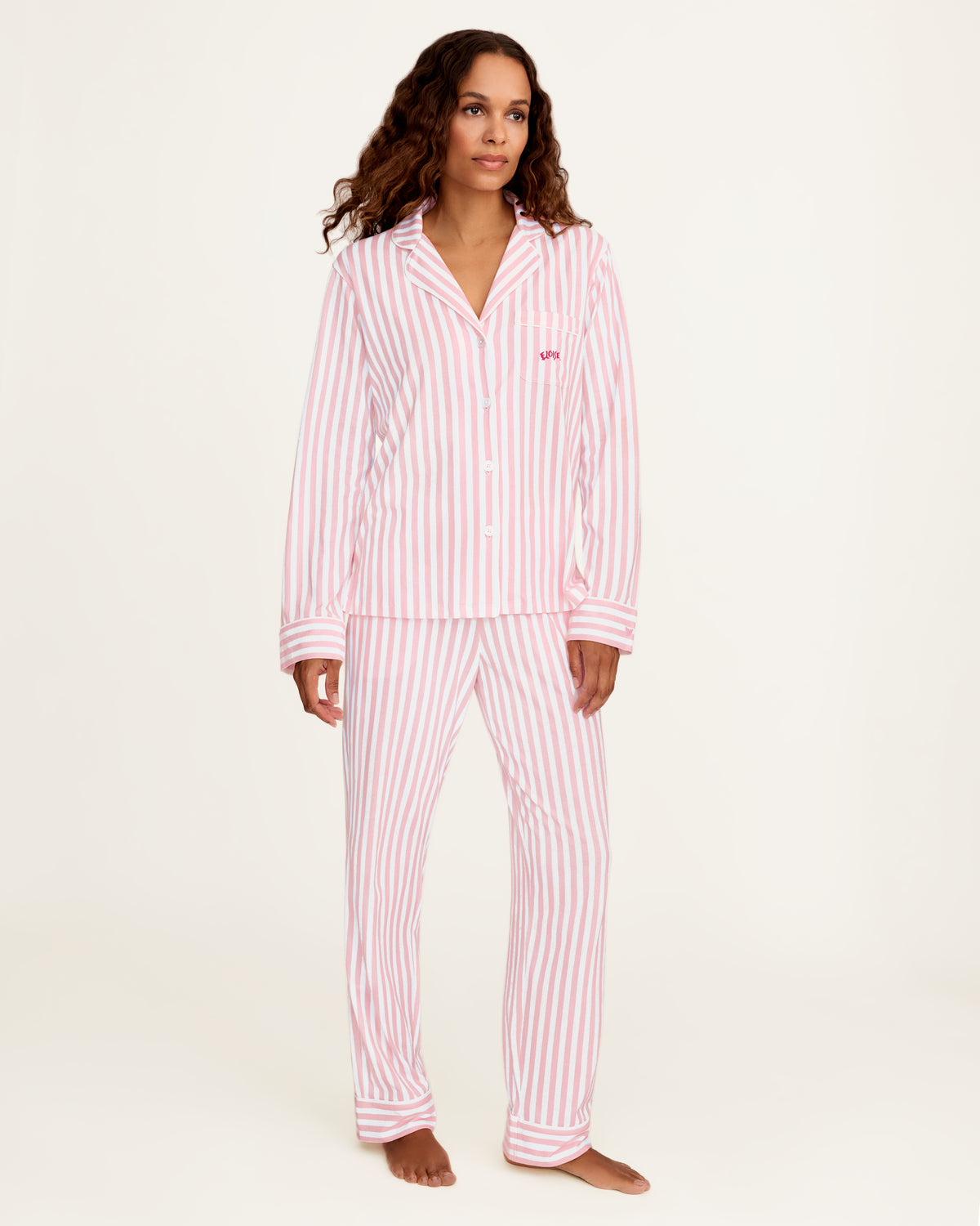A woman modeling a two-piece pajama set with pink and white vertical stripes, featuring a long-sleeve button-front top and matching pants, against a plain off-white studio background.