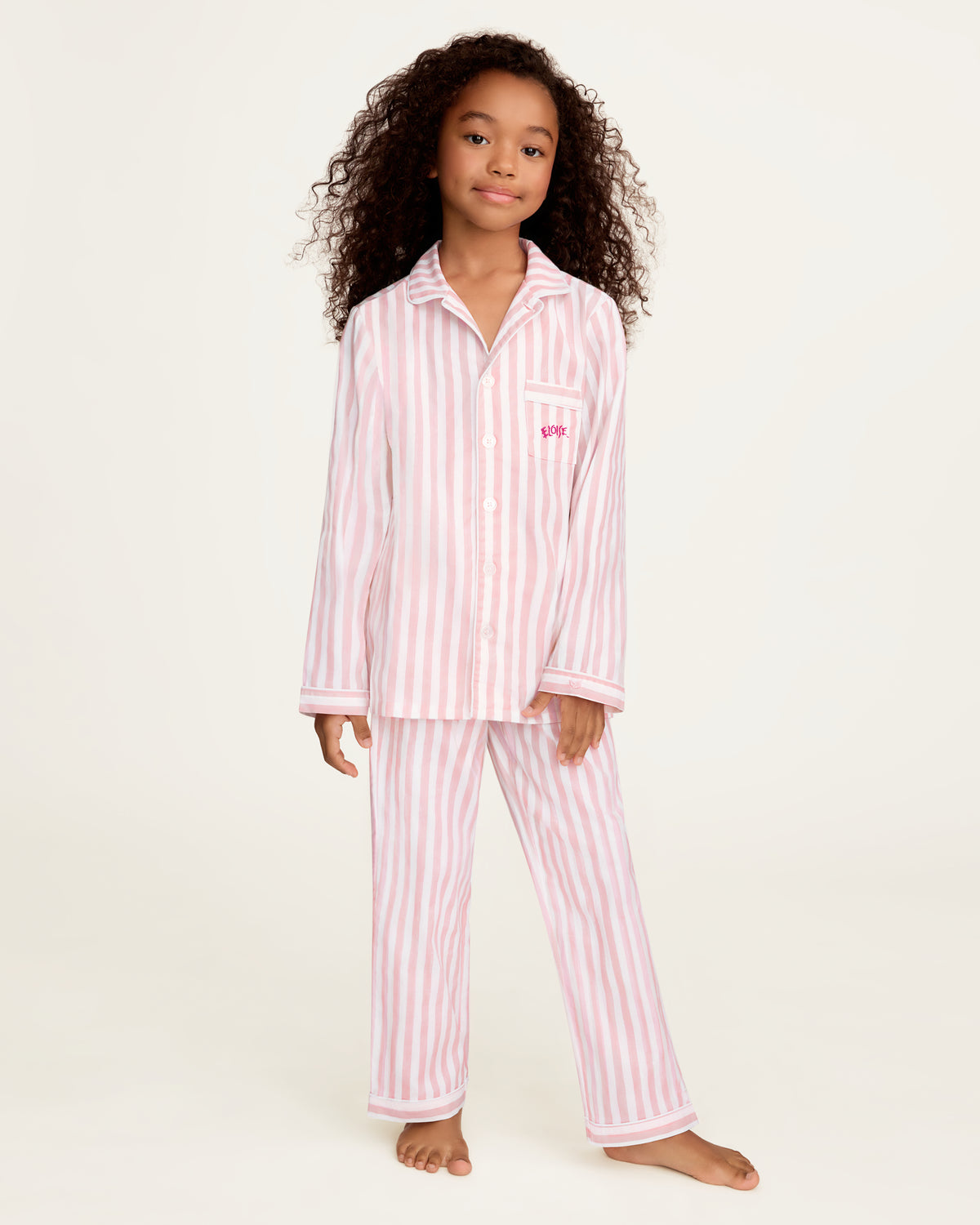 A young girl with curly brown hair stands barefoot wearing a pink and white vertically striped pajama set with a long-sleeve button-up top and matching pants, against a plain off-white studio background.