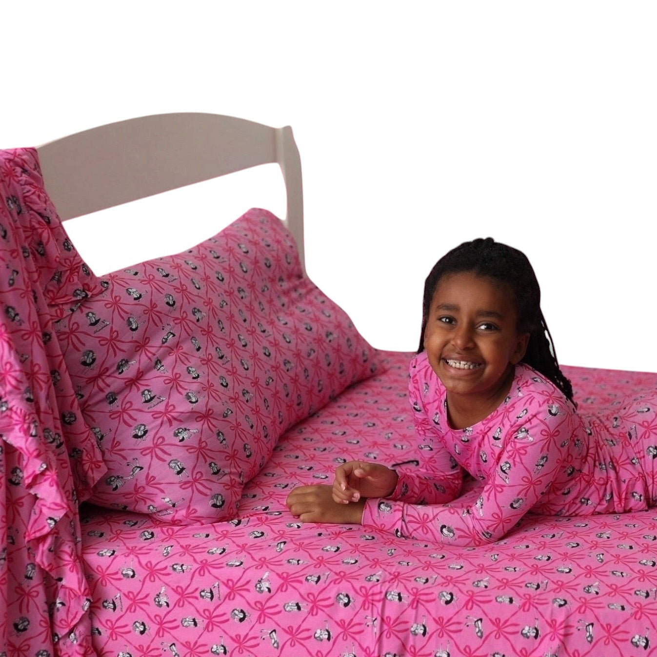 A smiling young girl lies on a bed  with a pink twin sheet set wearing a pink long-sleeve pajama set with a matching patterned pillow and bedspread. The pajamas and bedding feature a repeating black and white graphic pattern on a bright pink background.
