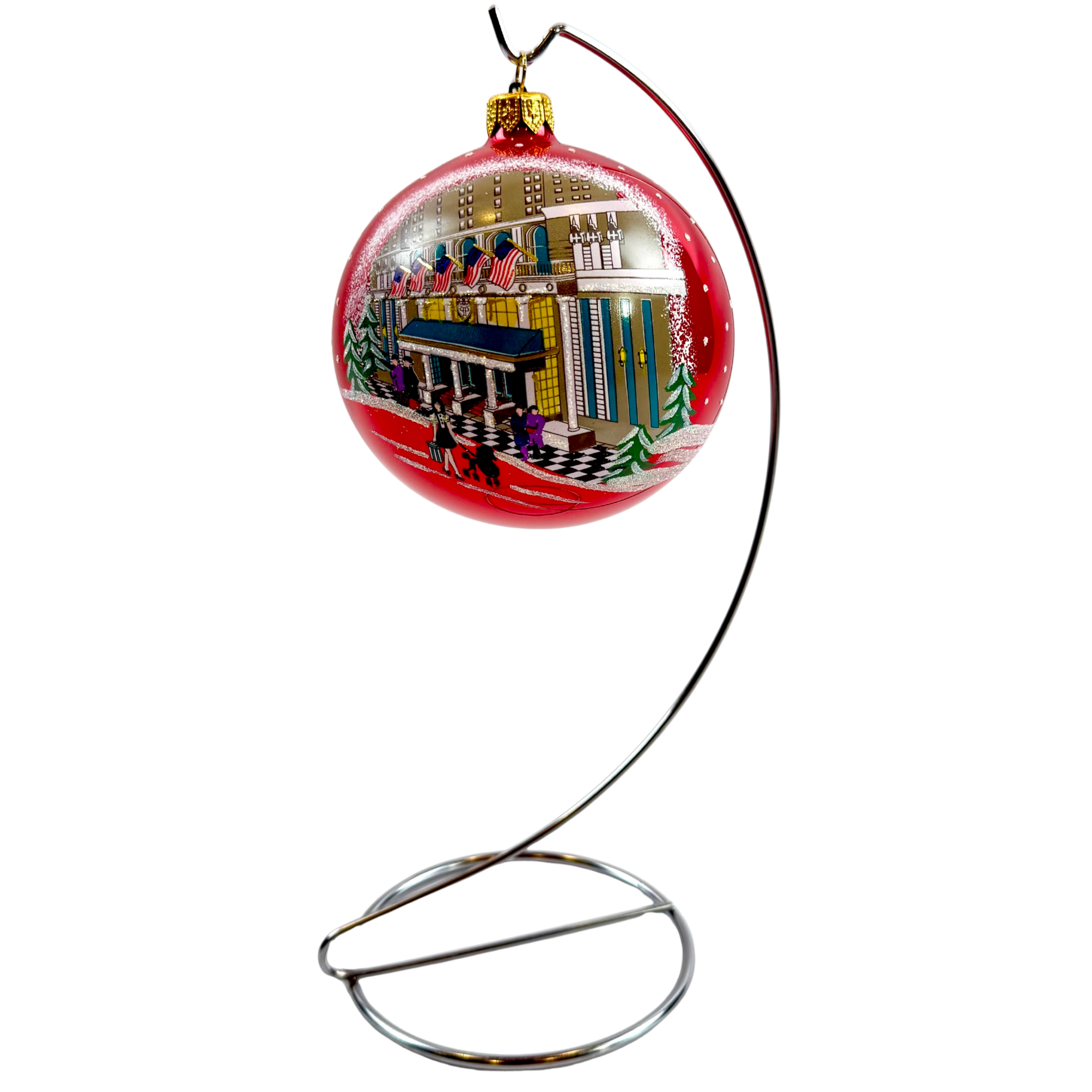 A red Christmas ornament displayed on a curved metal stand against a plain white background. The ornament features a hand-painted winter city scene with snow, trees, and colorful buildings.